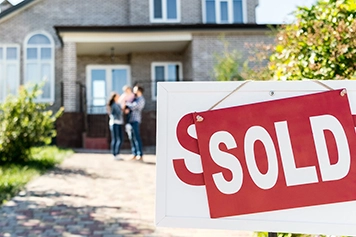 sold sign with couple holding child in background in front of house
